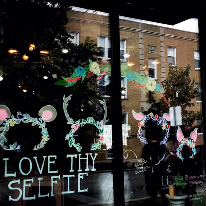 Colorful hand-drawn window installation reading Love Thy Selfie with floral animal ears.